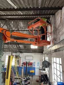 An orange scissor lift raised up inside a workshop with a worker standing on it, close to the corrugated metal roof. The workshop appears well-used with various tools and machinery around, including an engine hoist and a workbench.