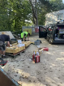 Workers preparing equipment and tools from a black van parked next to a tree and a building. The area is cluttered with construction materials and equipment.