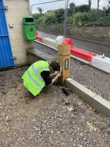 A worker in a green high-visibility jacket is installing or repairing a gate entry system, kneeling down near a blue metal gate.