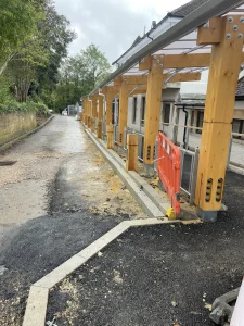 Construction of a covered walkway next to a building, featuring exposed wooden beams and construction barriers along a pathway.