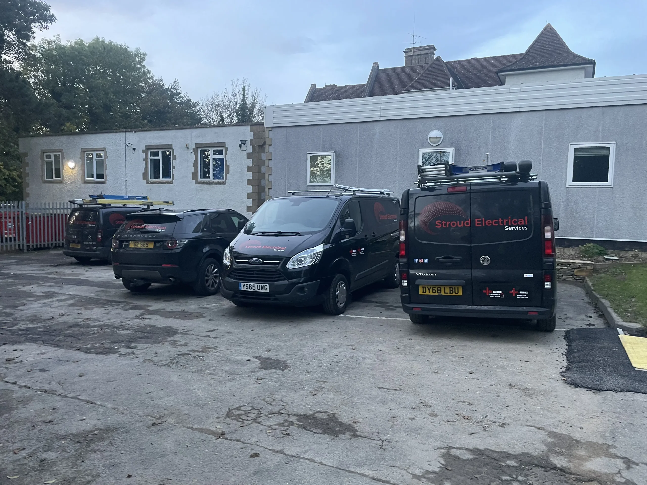 Evening view of a parking area with multiple vehicles, including vans marked "Stroud Electrical Services", parked outside a building.