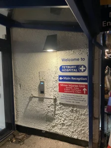 A sign at the entrance to Tetbury Hospital detailing the main reception and minor injuries unit, with an intercom system visible.